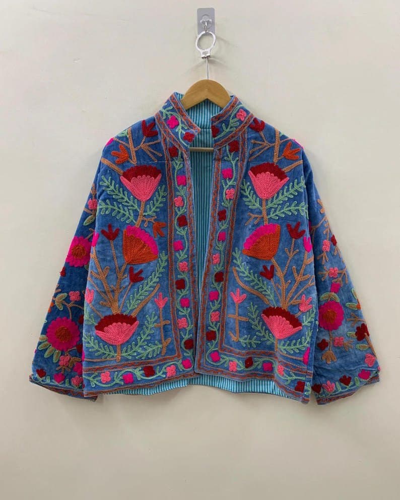 A cornflower blue Suzani jacket with pink and red floral embroidery, on a hanger against a pale wall