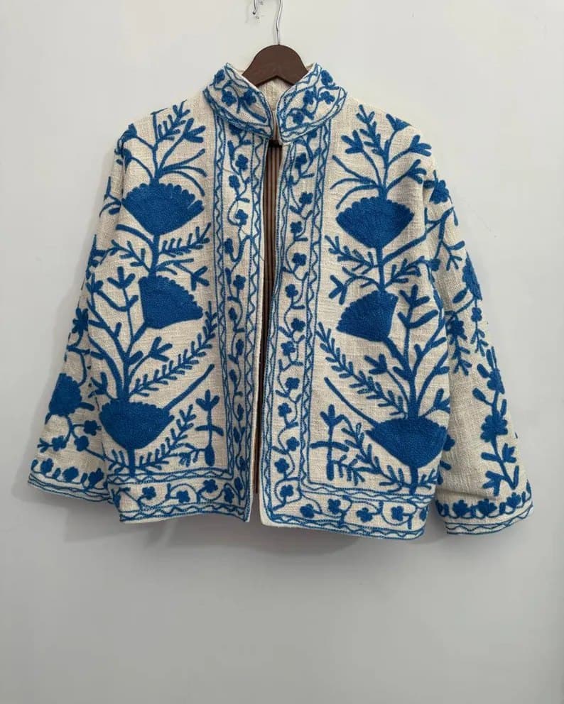 A ivory Suzani jacket with deep blue floral embroidery, on a hanger against a pale wall