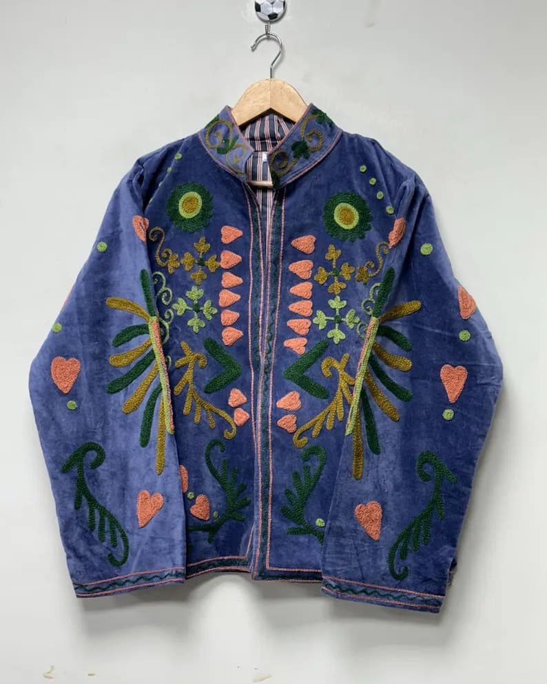 A periwinkle velvet Suzani jacket with coral and green motifs, on a hanger against a pale wall